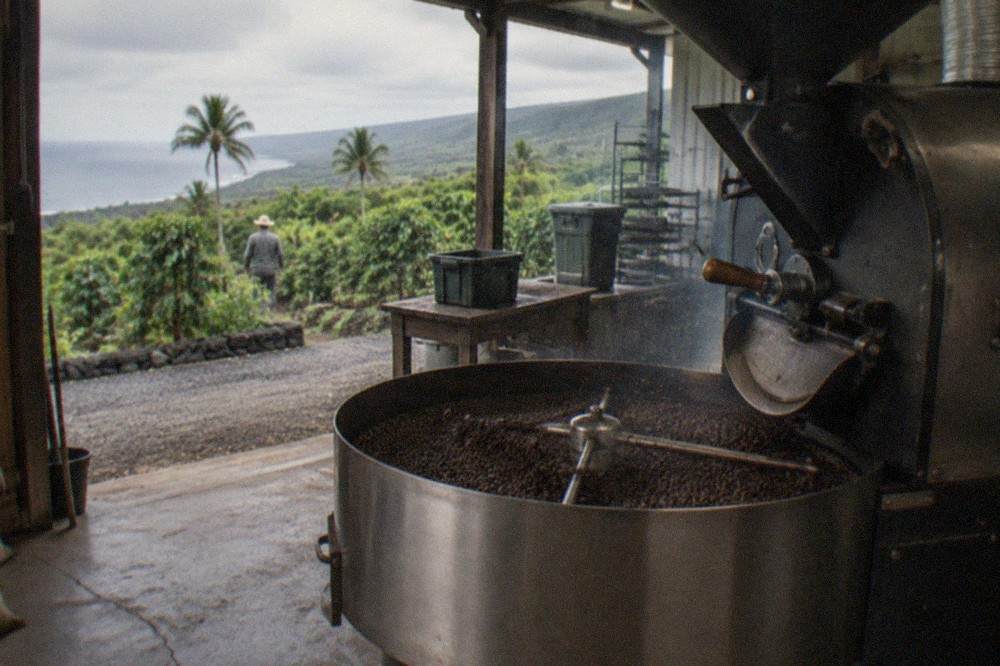 Roasted Kona coffee beans cooling in a metal drum beside a farm roaster